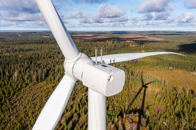 Ett stort vitt vindkraftverk står högt ovanför ett skogslandskap, med blå himmel och spridda moln i bakgrunden. Turbinens blad visas tydligt och kastar skuggor i terrängen nedanför.